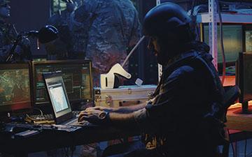 Man wearing combat gear sitting at a small laptop, utilizing Viasat services in the field